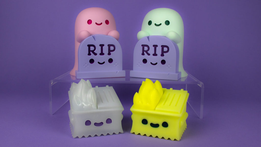 The Lil' Ghosty Night Light and its pink variant pictured behind the Dumpster Fire Ghost and its Glow in the Dark variant