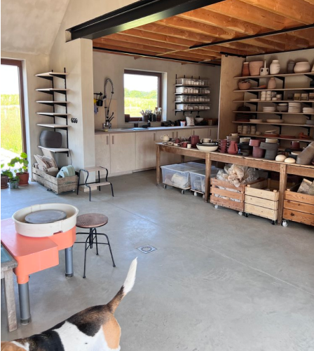 The interior of the ceramics studio in a rustic building with a pottery wheel on theleft and stacks of complete and incomplete hand-thrown plates and vessels