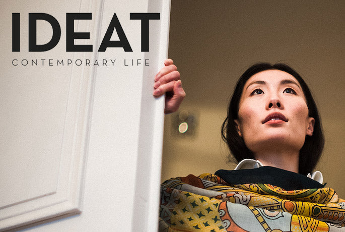 IDEAT logo superimposed over a large white open shutter, a woman looks out from the window having just opened it. She is wearing a luxury scarf from Alba Amicorum