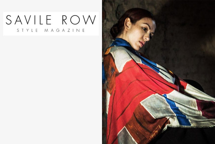 Savile Row style magazine text logo on the left, woman seated wearing a Man Ray Alba Amicorum luxury scarf in reds and blues. She is in front of dark brown textured wall.