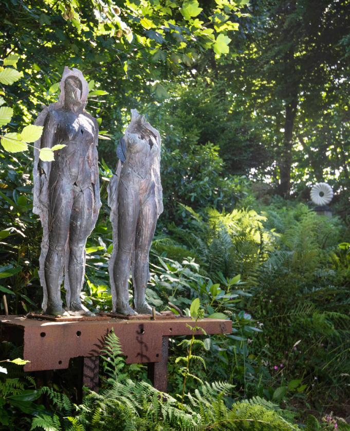 two stone figurative standing sculptures in a lush green garden mounted on a wooden beam