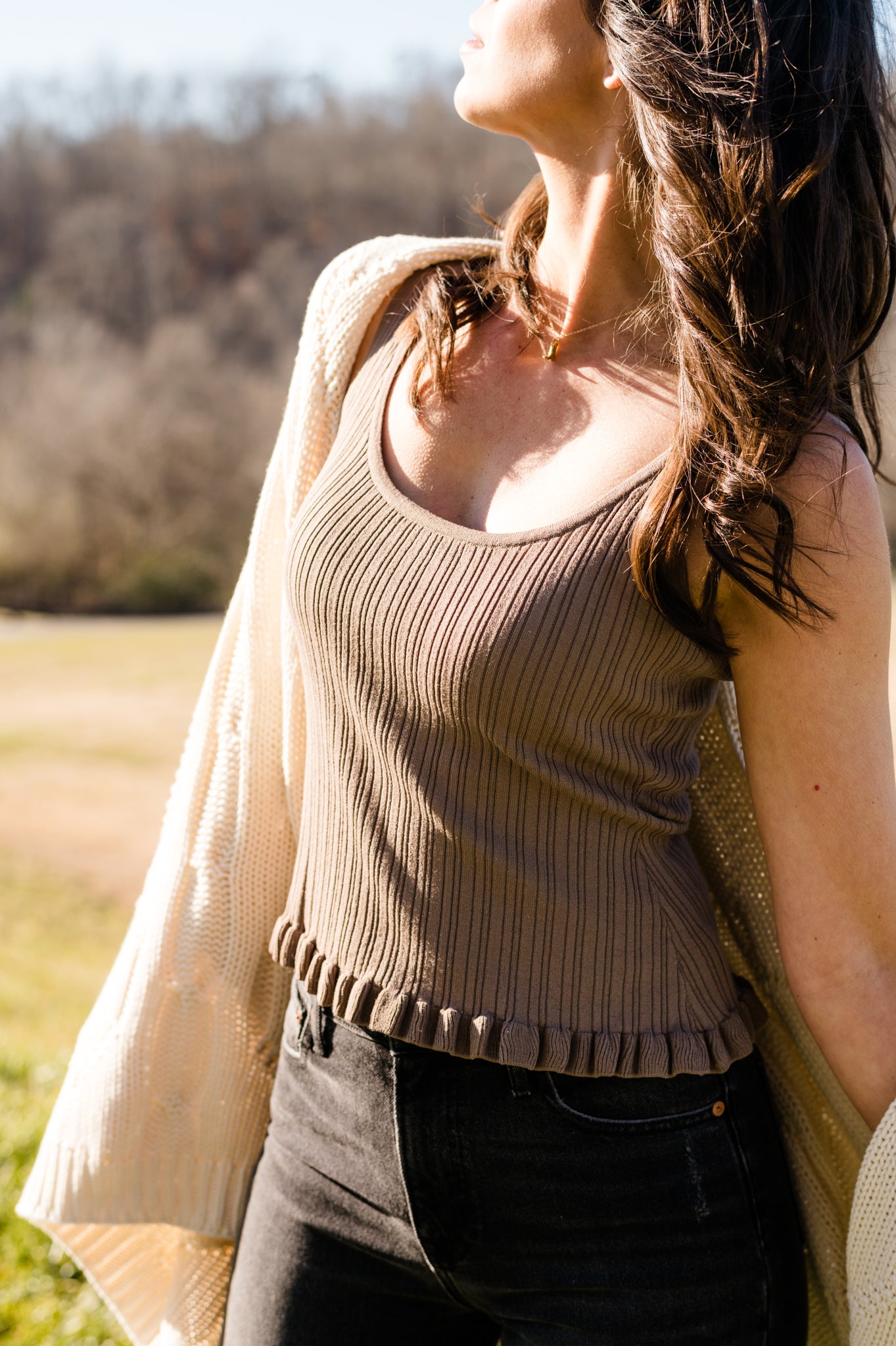 Woman in beige top and black jeans, with sunlit nature backdrop.