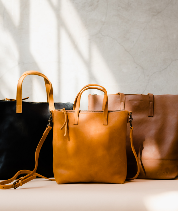 Three leather handbags in black, mustard, and brown colors against a light background with shadows.