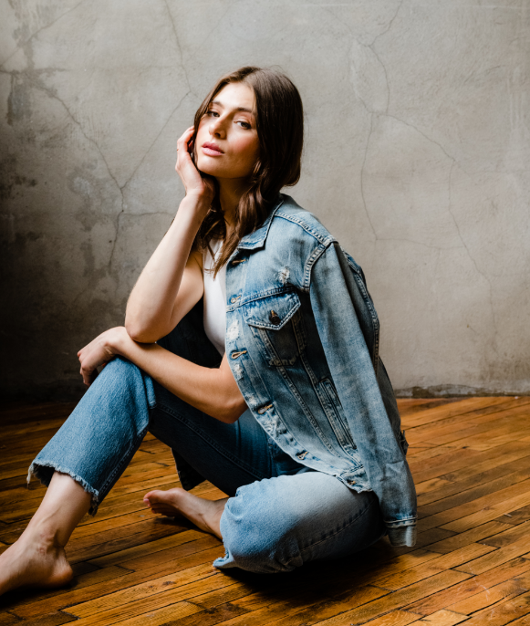 Woman in denim outfit sitting on wooden floor against a textured wall.