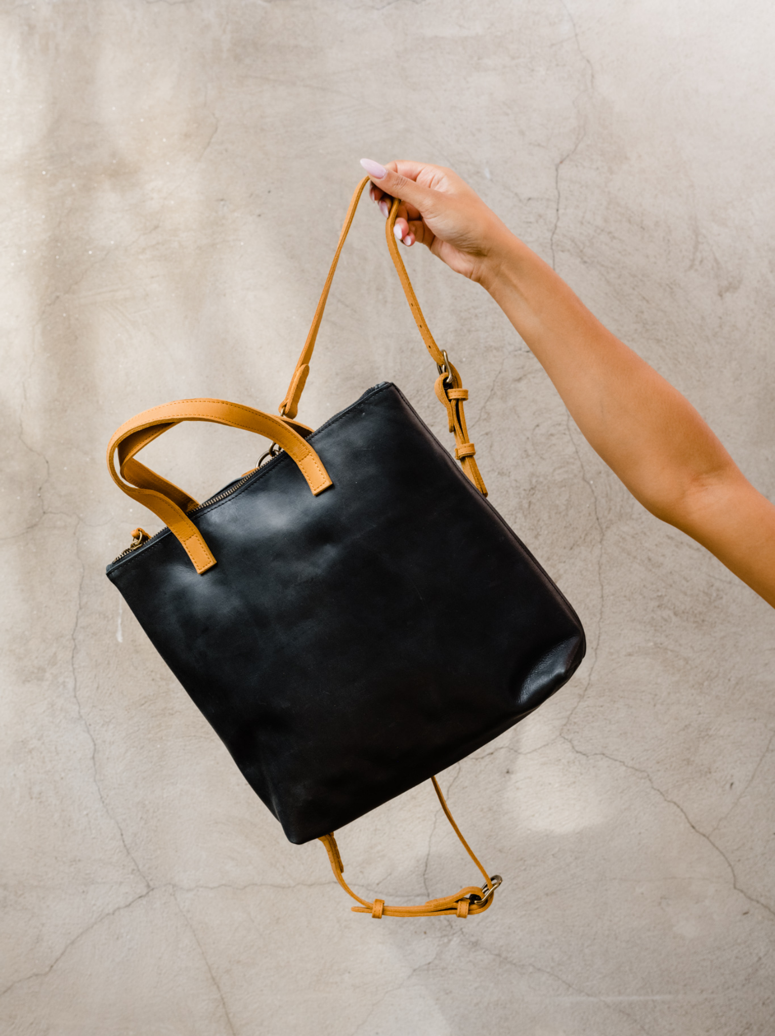 A person's hand holding a black leather bag with a tan strap against a textured background.