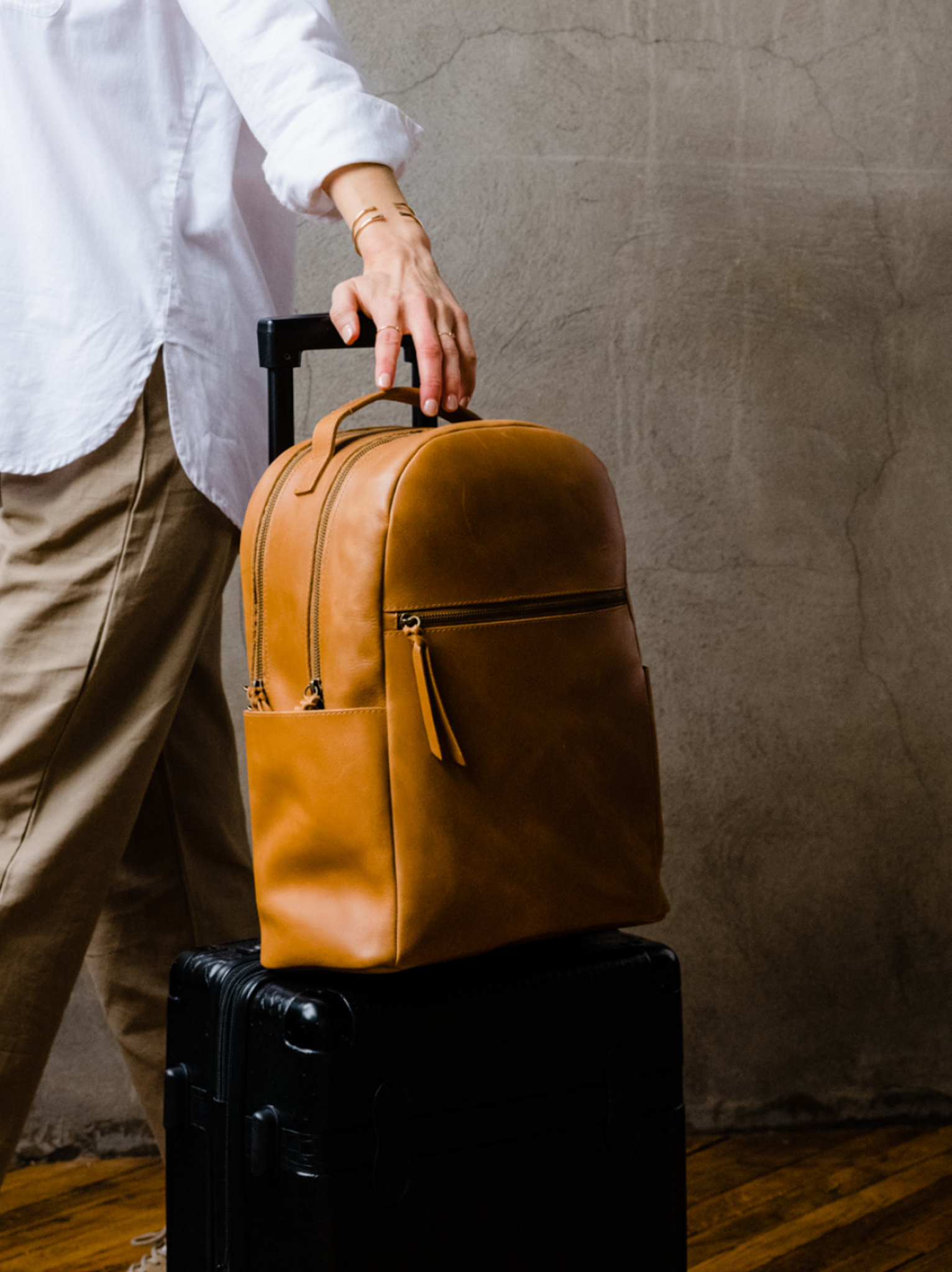 Person resting hand on a brown leather backpack atop a black suitcase against a textured wall.