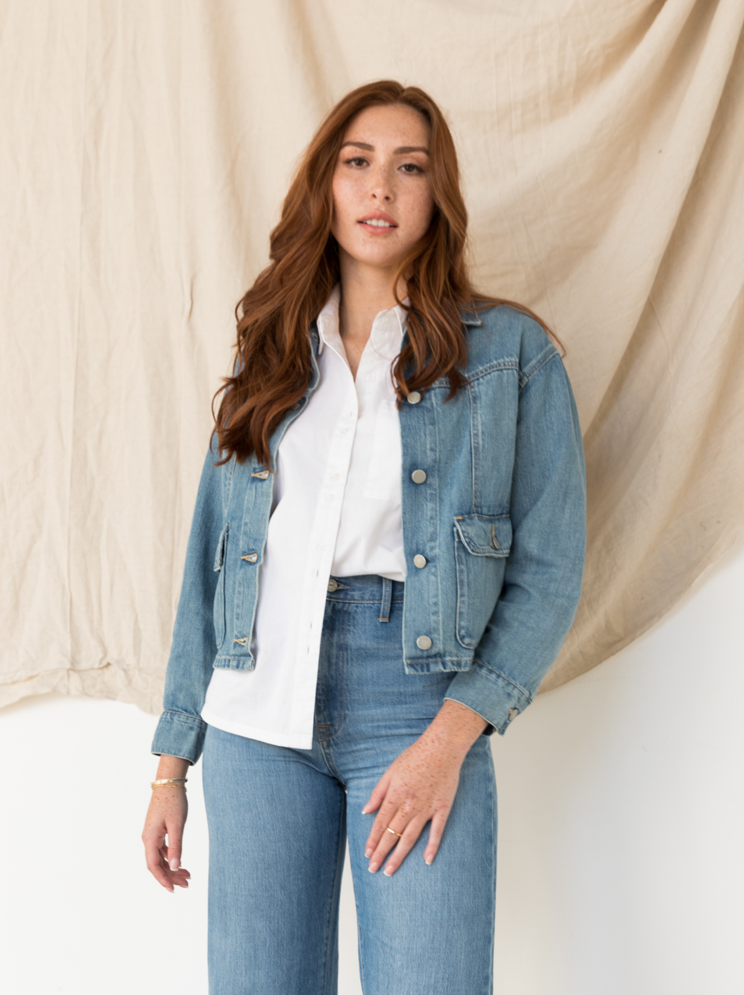 Woman in denim jacket and jeans standing against a beige backdrop.