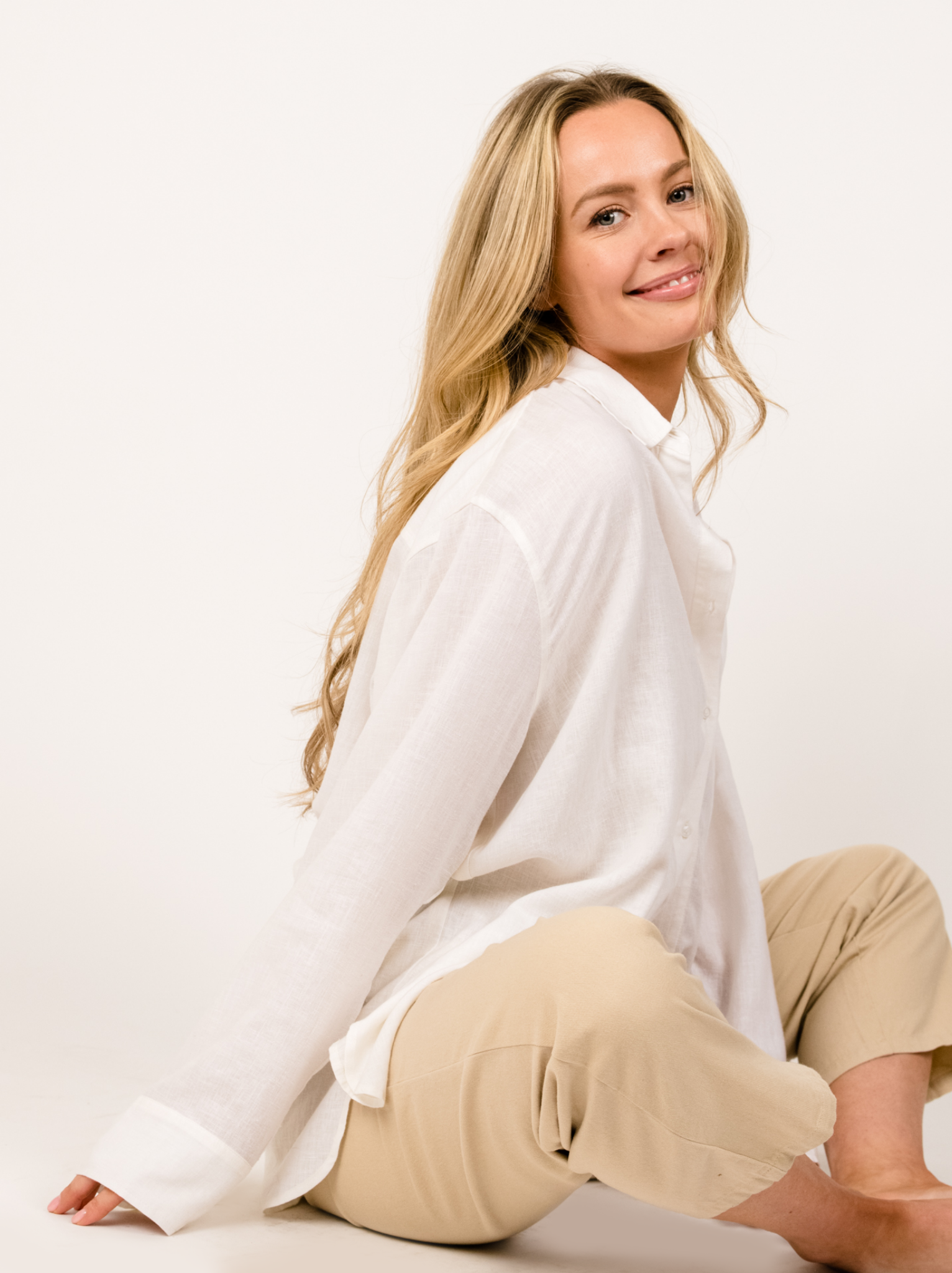Woman in white shirt and beige pants sitting against a white background.