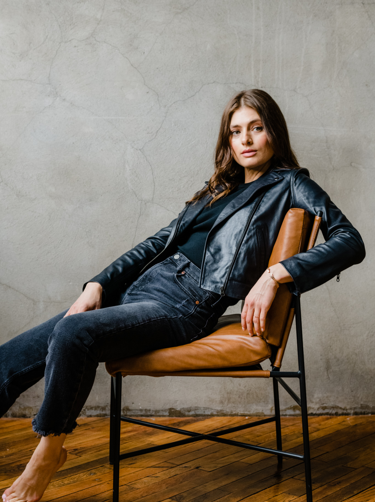Woman sitting casually in a chair wearing a black leather jacket and jeans.