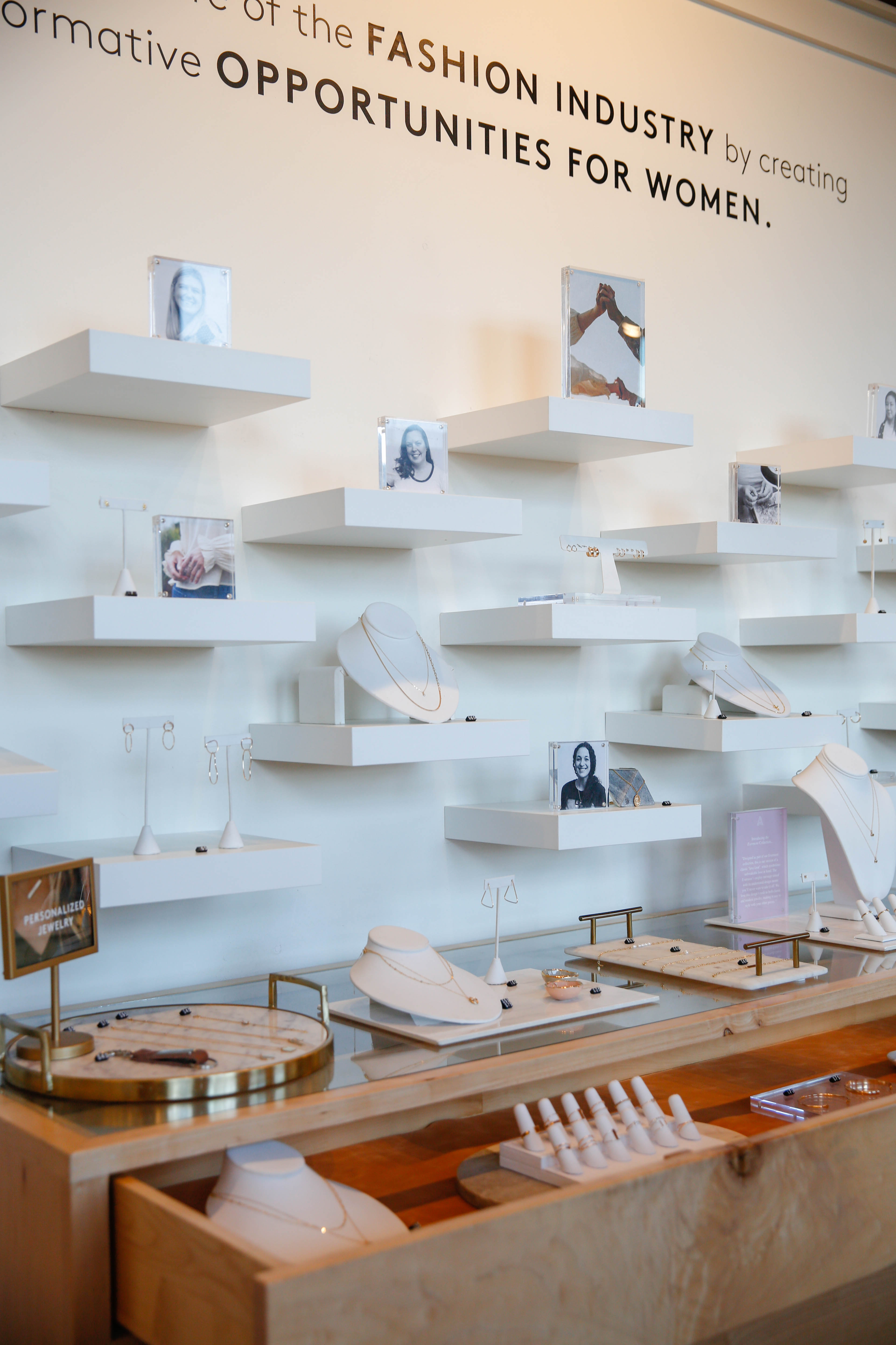 Jewelry shop display with necklaces, rings, and photographs on white floating shelves and a wooden counter.