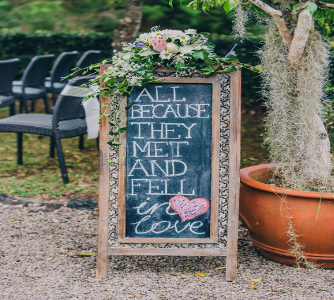 A sign that says “All because they met and fell in love” is displayed on a chalkboard sign with flowers and a pink heart. 