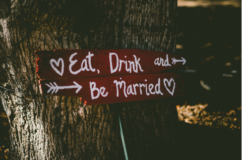 A sign made of wooden boards displayed outdoors reads “Eat, Drink, and Be Married” with hearts and arrows.