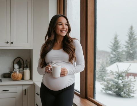 Young pregnant woman holding cup of tea and looking at falling snow