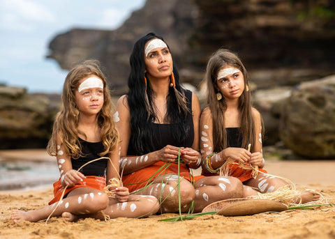 Tegan Murdock sits at the beach in traditional aboriginal paint and clothing with her two daughters weaving with grass