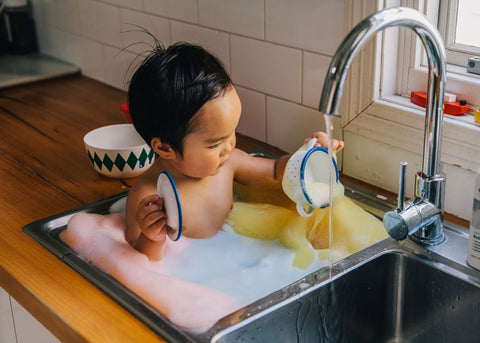 photo of a baby boy with black hair in a kitchen sink bath filled with rainbow bubbles