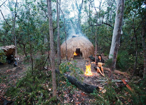 Photo of Claire Dunn by her campfire with her handmade hut behind her in the Australian bush