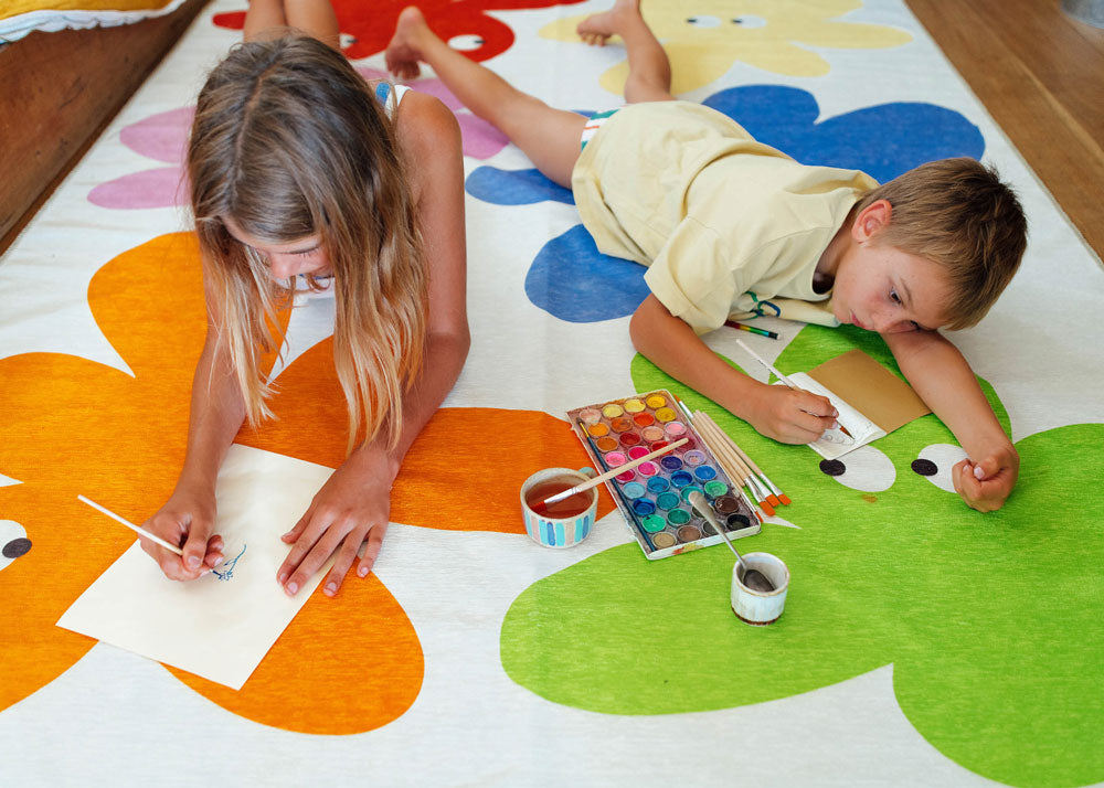 two young children lie on their stomachs on a rug painting with watercolours