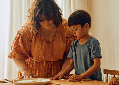 A photo of Julia Ostro with her child making pasta. She wears an orange dress and he has a blue tshirt and short black hair.