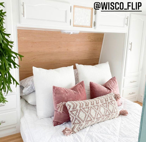 A close up image of an RV bed with a natural wood built-in headboard, white cabinets hanging above, Beddy's Love at First White Beddy's on the floor-style bed, with mauve pink accent pillows.