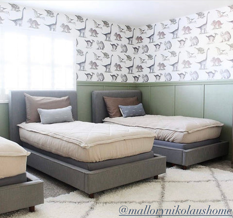 Three twin beds sit in a bedroom with board and batten half way up the surrounding walls painted olive green and a multi colored dinosaur print wallpaper goes up the remaining top half of the wall. Beds all feature our oatmeal colored Naturally Boho Beddy's.