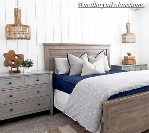 A dark brown queen bedframe sits in front of a white vertical striped shiplap wall. Gray nightstands sit to each side of the bed and wicker coastal style hanging light pendants hang above each one. Bed features our Nautical Navy Beddy's. 