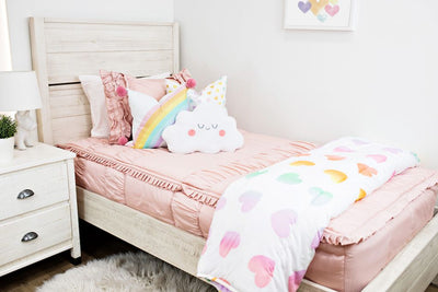 twin bed with pink ruffle bedding and white pillow with rainbow ombre hearts, white pillow with rainbow and pink pom poms on the corner, plush marshmallow pillow, and white blanket with ombre hearts at the foot of the bed