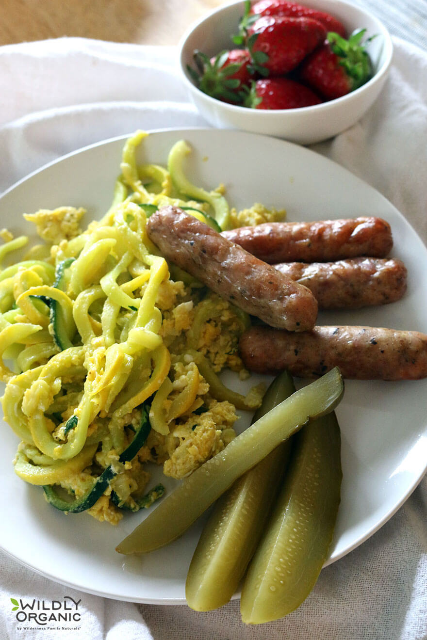 shite plate with squash and zucchini noodles, sausage, sliced pickles and bowl of strawberries