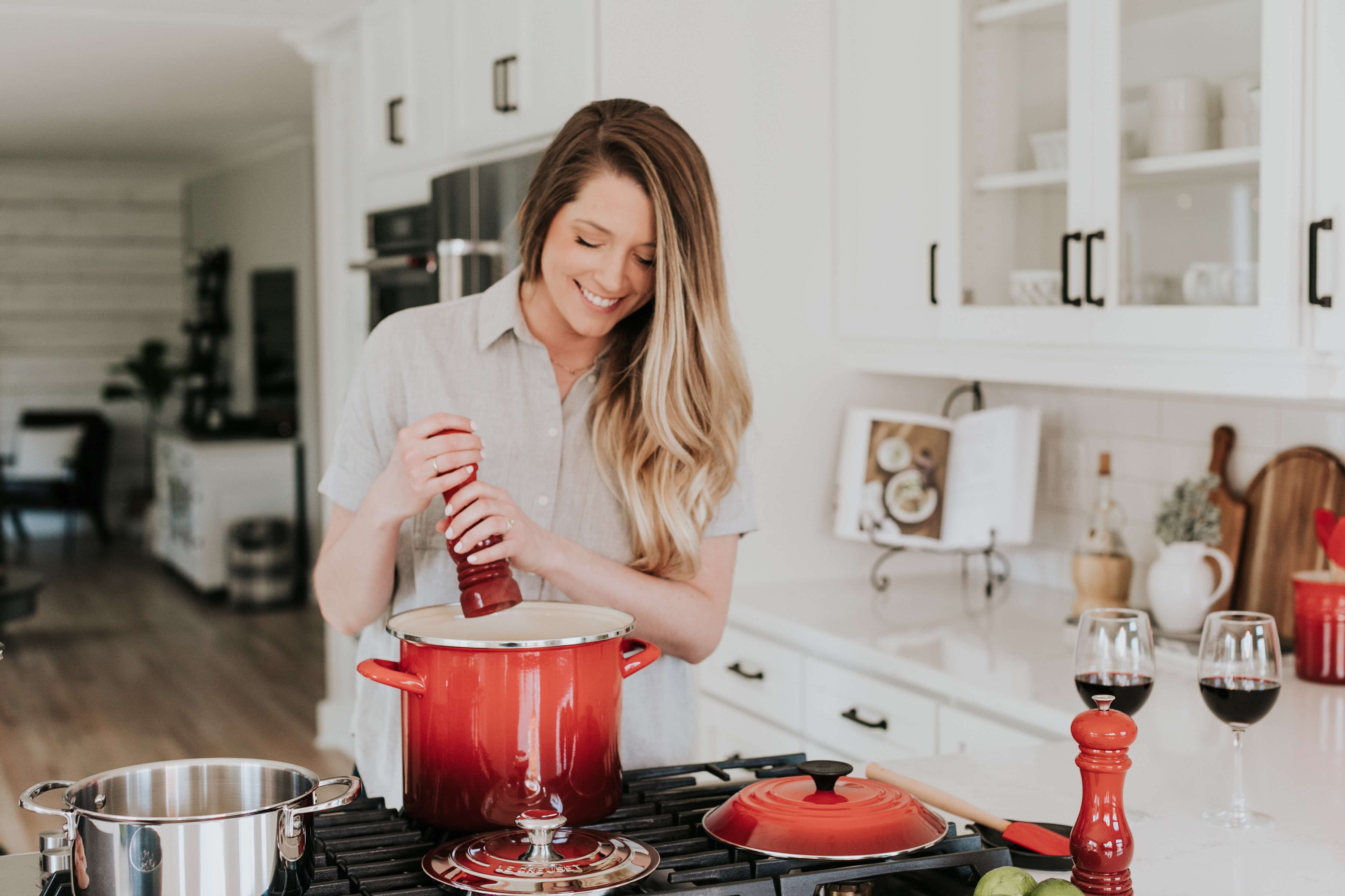 A young Blonde woman smiling and sprinkling black pepper into  a red pot on a stove