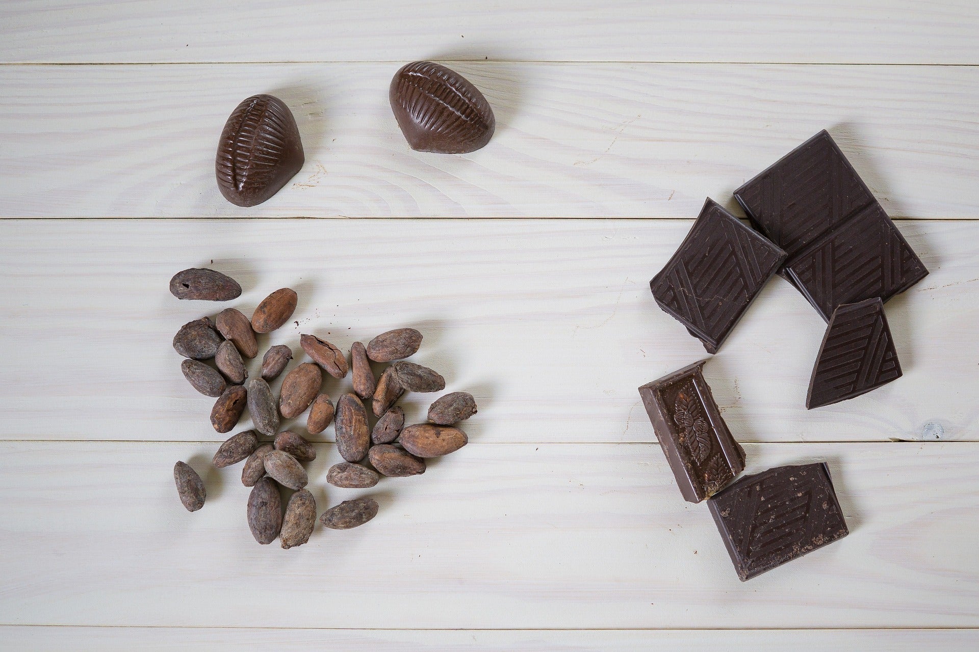 Cocoa beans and broken pieces of dark chocolate on a white wooden surface