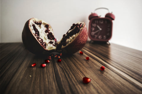 A photo of a split pomegranate on a wooded tabletop and a wind up alarm clock in the background.