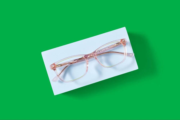 A pair of pink rectangular eyeglass frames sitting on a blue block on top of a green background.