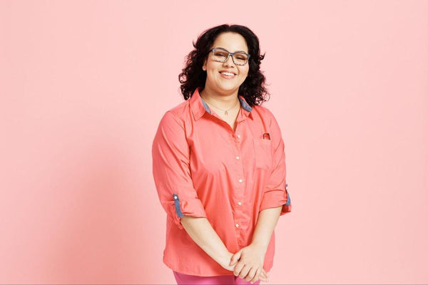 Woman smiling wearing glasses and a pink shirt in front of a pink background.