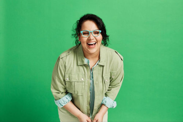 Laughing woman with curly hair wearing teal round eyeglass frames in front of a green background.