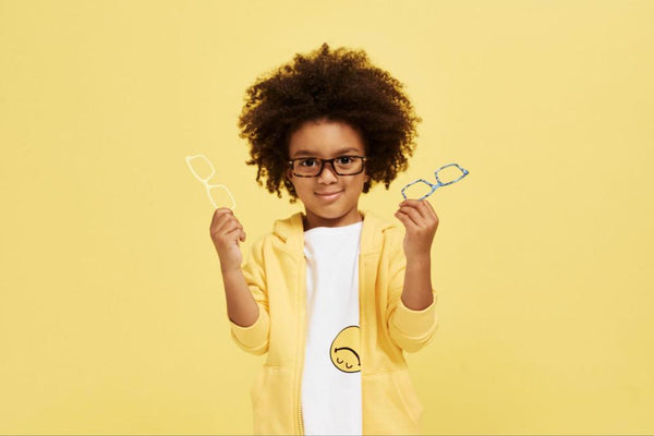 A male child wearing boys' glasses in front of a yellow background and holding two pairs of glasses.