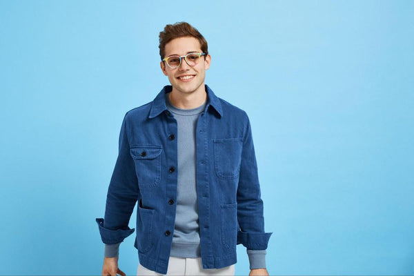 A young man wearing a blue jacket and colorful round eyeglass frames in front of a blue background.