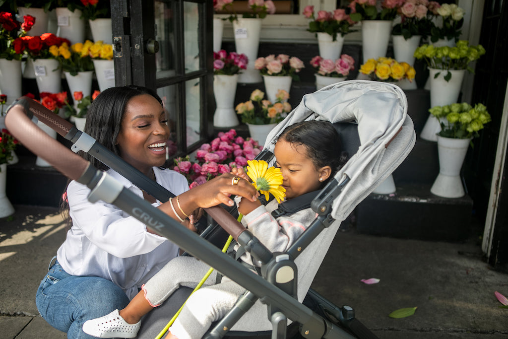 Mom Kneeling Beside Her Daughter in A UPPAbaby CRUZ V2 Letting Her Smell A Flower