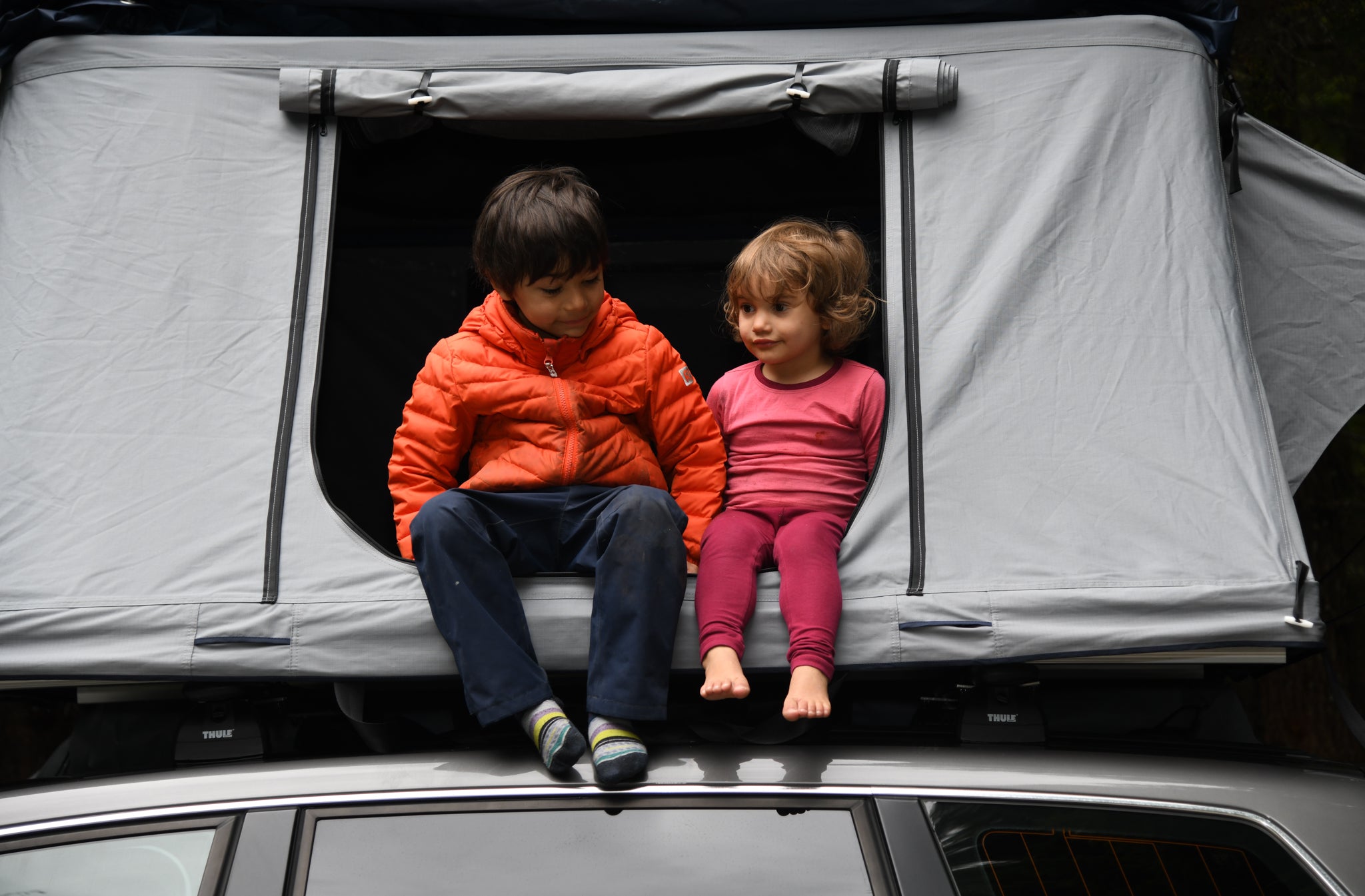 A young boy and girl sit in a roof top tent on a camping trip