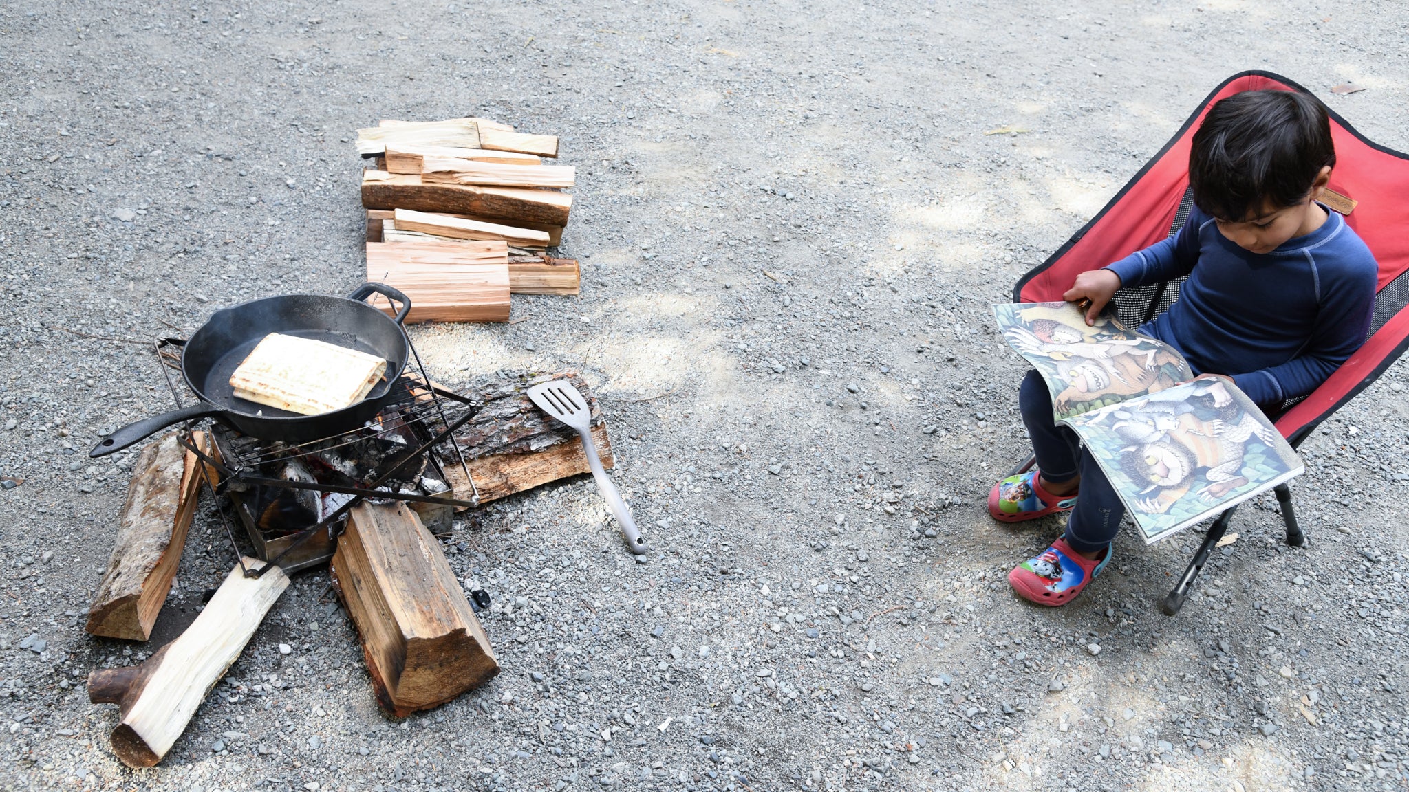 Young boy reading in a red camp chair beside a small campfire where a grilled cheese cooks in a cast iron pan.
