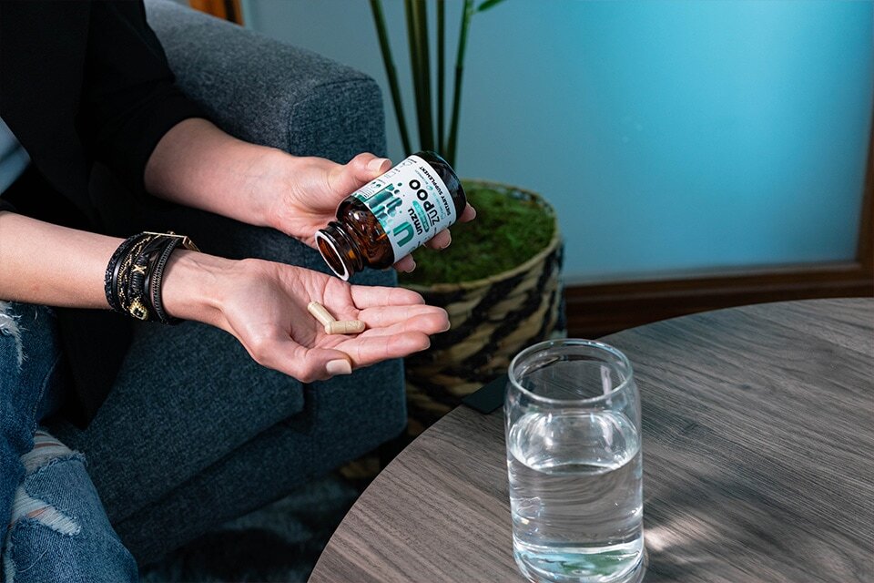 Woman holding a bottle of zuPOO sitting on a sofa next to a wooden table.