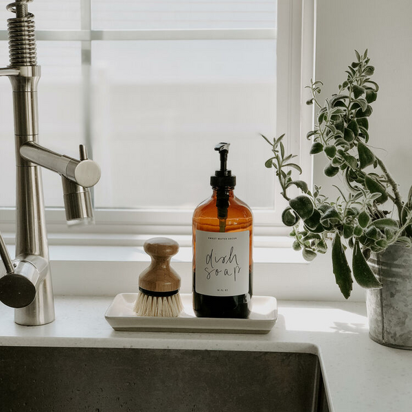 Amber jar dispenser bottle and a kitchen brush scrubber styled on a decorative ceramic tray on a kitchen sink.