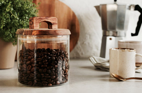 A glass jar of coffee beans, with a mug, spoon and stovetop coffee maker next to it.