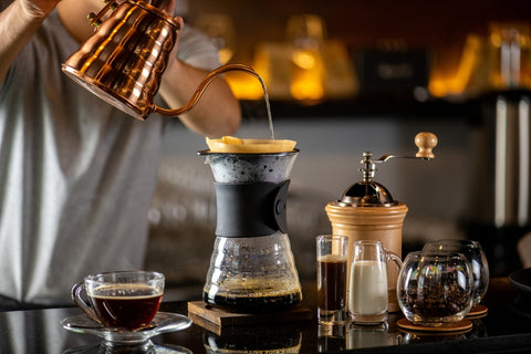 Man pouring hot water from a kettle into a pour-over coffee maker, with a cup of coffee, grinder and sugar jar next to it.