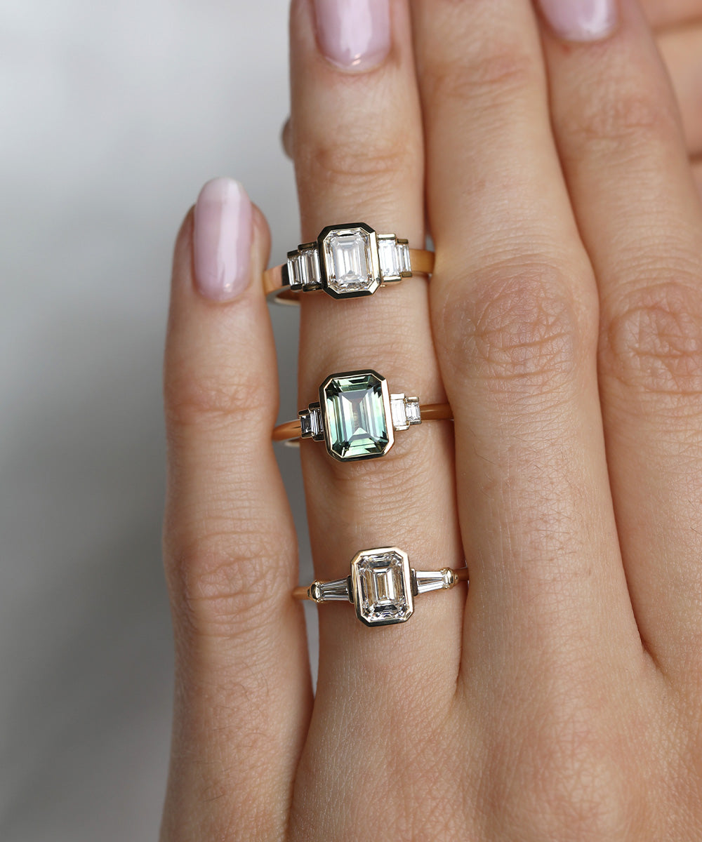 Three Art Deco-inspired alternative emerald cut engagement rings on the hand with full rubover bezel settings