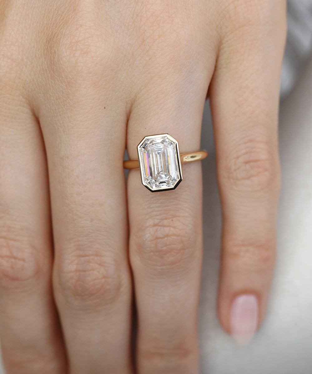 3ct natural emerald cut diamond engagement ring with a full bezel rubover setting in 18ct yellow gold and a delicate d shape band.