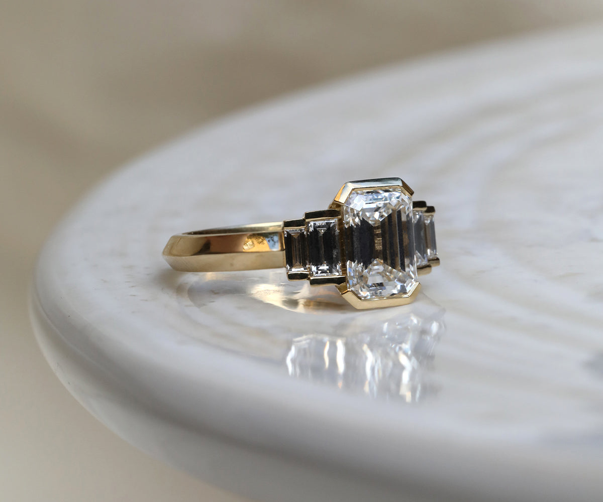 Art Deco-inspired emerald cut engagement ring with a half bezel rubover setting and stepped baguette cut diamonds and 18ct yellow gold band.