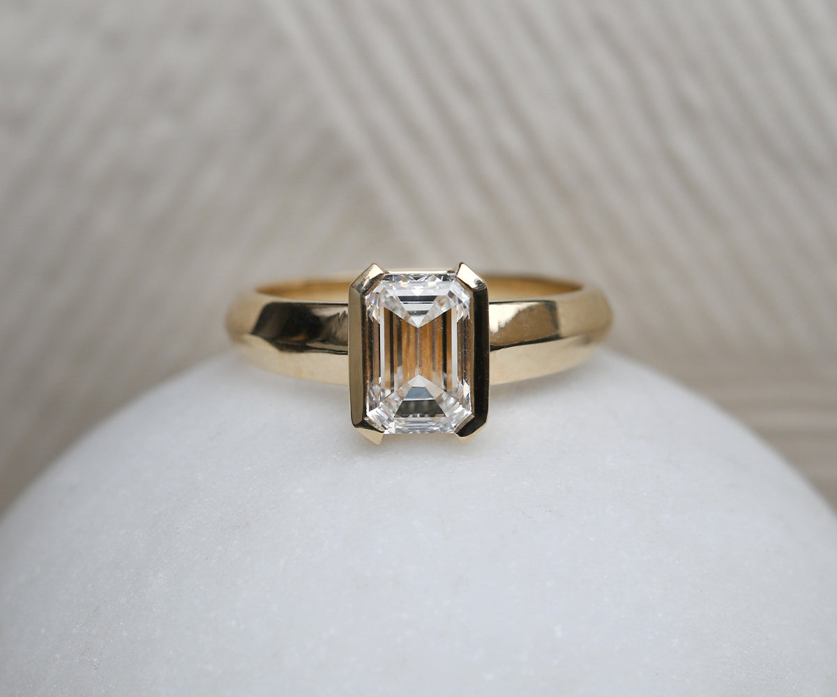 Lab grown emerald cut diamond engagement ring with a half bezel rubover setting and statement chunky 18ct yellow gold tapered band.