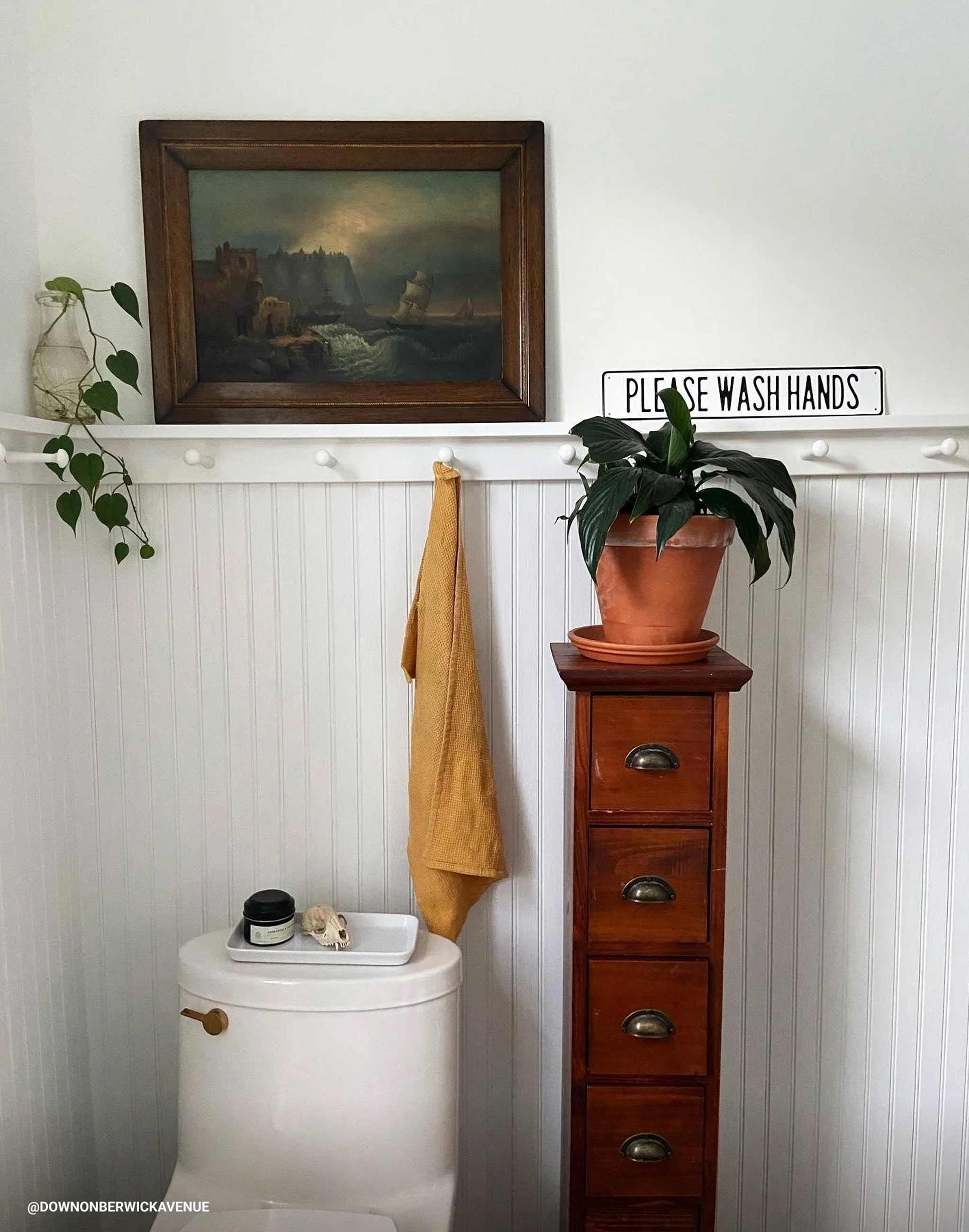 White bead-board behind toilet in bathroom with potted plants surrounding it. 