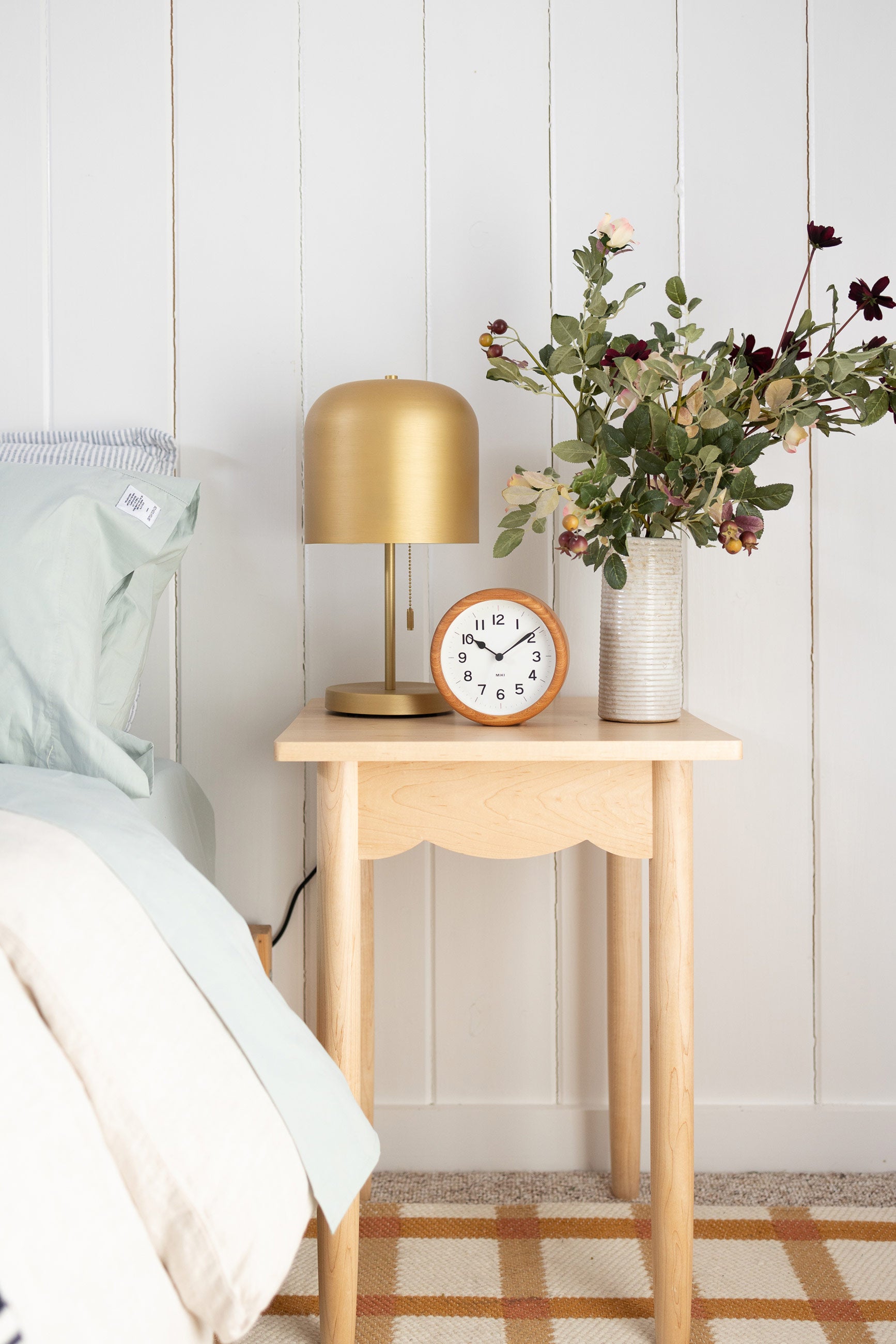 Shaker-style bedside table with brass lamp, small wooden clock, and fresh cut flowers in a white vase