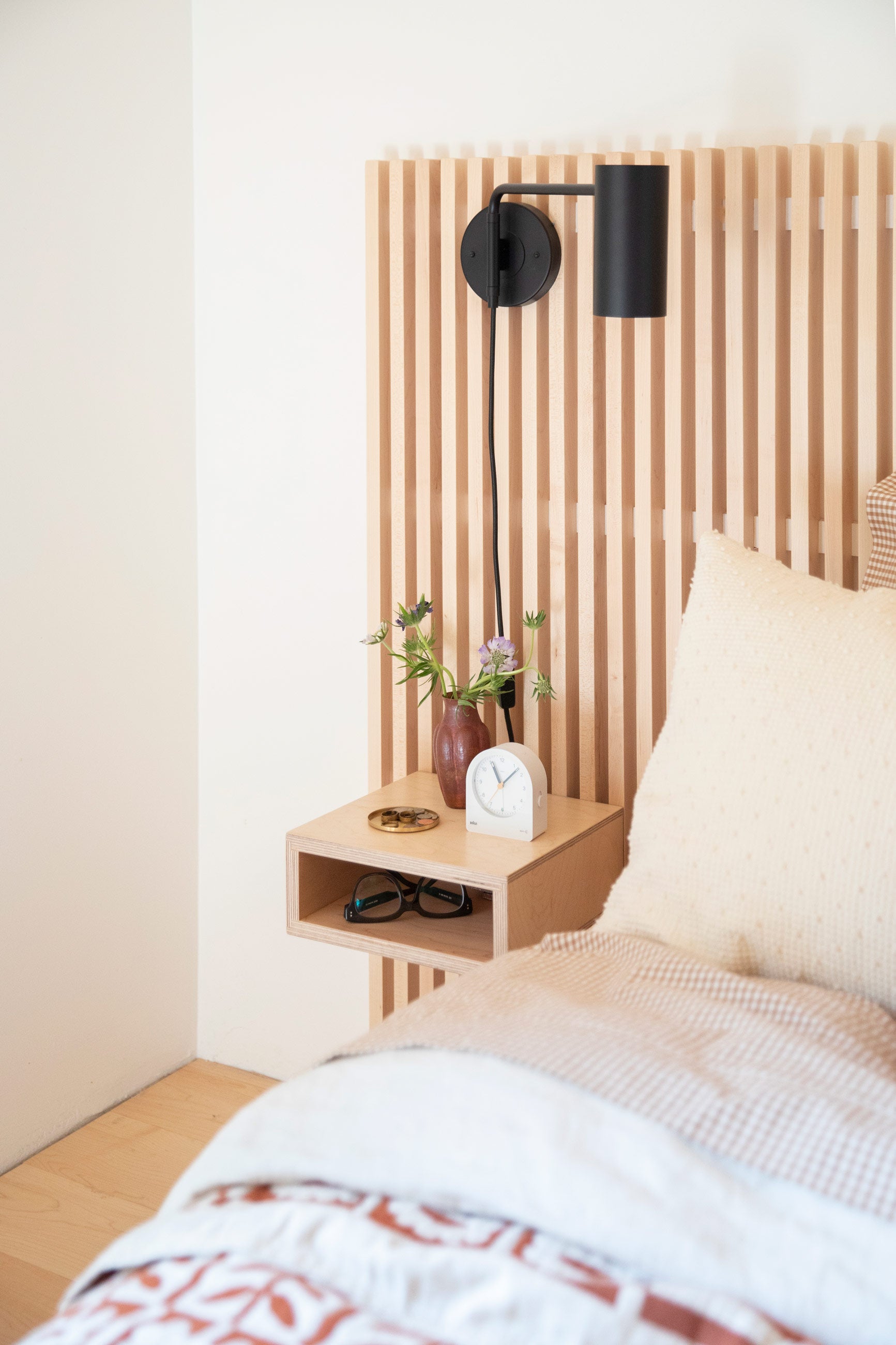 Bedside table with small white clock, vase full of fresh flowers, and black wall sconce.