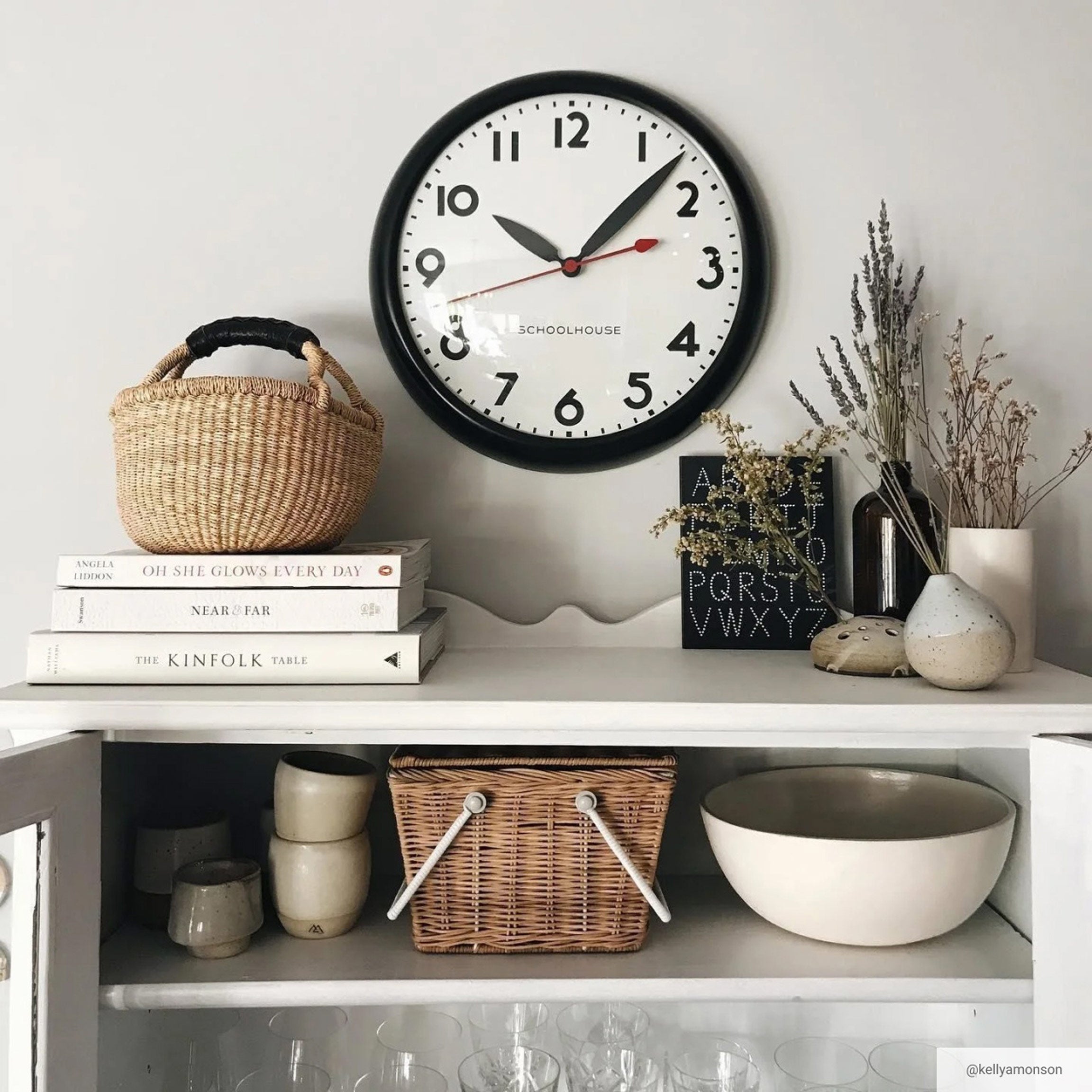 Black wall clock above white cabinet holding baskets, dish ware, books, and greenery.